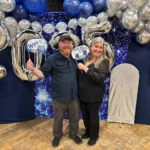 Couple posing happily in front of blue and silver balloon decoration at a celebration