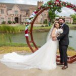 Bride and groom posing by lakeside under circular floral arch at Castle Waterford wedding