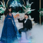 Couple dancing during an elegant engagement or wedding celebration while surrounded by white floral décor, chandeliers, and low-lying fog effects. The woman wears a sparkling royal blue gown, and the man is dressed in a black tuxedo as he kneels and holds her hand on a glowing dance floor.