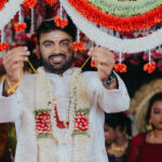 Indian groom dressed in a white sherwani and floral garlands smiling during a traditional wedding ceremony while holding decorative floral strings beneath vibrant red and white wedding decorations. Family members and guests are visible in the background.
