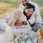 Romantic couple embracing during an outdoor picnic date on a blanket in a grassy park. The woman in a floral dress smiles while the man hugs her lovingly from behind, surrounded by picnic décor, flowers, and soft natural sunlight.