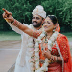 Traditional Indian bride and groom standing outdoors in wedding attire, smiling and pointing into the distance. The groom wears a white sherwani with a matching turban, while the bride is dressed in a red-orange bridal saree with heavy gold jewelry and floral garlands.