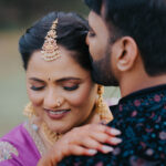 Traditional Indian couple sharing an intimate moment during a wedding photoshoot. The bride, dressed in a purple saree with elaborate gold jewelry and bridal makeup, smiles softly while the groom leans in close wearing a dark embroidered outfit.