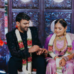 Indian bride and groom seated together during a traditional wedding ceremony while exchanging rings and smiling warmly. The bride wears a purple saree with layered gold jewelry, and the groom is dressed in a black embroidered sherwani. Both wear floral garlands against an ornate decorative backdrop with vibrant floral accents.