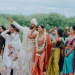 Indian wedding couple standing with family and friends outdoors during a joyful celebration. The bride and groom, dressed in traditional attire with floral garlands, smile and point into the distance while guests in colorful sarees and ethnic outfits celebrate around them.