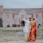 Indian bride and groom standing together outdoors in front of a grand castle-style wedding venue. The groom, dressed in a white sherwani with floral garlands, points into the distance while the bride in a red-orange saree smiles beside him near a scenic lakeside setting.