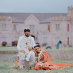 Indian bride and groom posing outdoors in front of a grand castle-style venue. The groom, dressed in a white sherwani with floral garlands, sits beside the bride in an orange-red bridal saree as she rests her head on his lap in a romantic moment near a lakeside lawn.