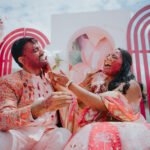 Indian bride and groom laughing joyfully during a colorful pre-wedding celebration while applying red powder on each other’s faces. The couple is dressed in vibrant traditional attire and seated in front of a decorative pink and floral backdrop under bright daylight.