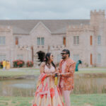 Indian couple standing together outdoors in front of a grand castle-style wedding venue. The couple is dressed in colorful traditional wedding attire and smiling at each other beside a scenic pond and landscaped greenery.