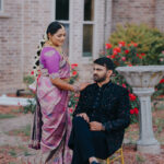 Traditional Indian couple posing outdoors in a garden setting near a brick mansion. The bride is wearing a pink silk saree with gold jewelry and floral hair accessories, while the groom sits on a chair in a black embroidered sherwani as they share a romantic moment.