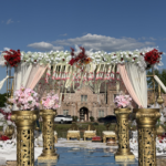 Luxurious outdoor wedding mandap setup featuring a mirrored aisle lined with ornate gold floral pillars and abundant white, red, and pink floral arrangements. Elegant drapes and hanging floral décor frame the ceremony stage against a bright blue sky and castle-style venue backdrop.