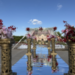 Luxury outdoor wedding aisle featuring a mirrored walkway lined with ornate gold floral stands and vibrant pink, red, and white flower arrangements leading toward an elegant floral mandap setup under a bright blue sky.