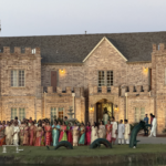 Large group of wedding guests gathered outdoors in front of a grand castle-style venue during an evening celebration. Guests dressed in colorful traditional attire stand near a lakeside lawn with elegant architecture and warm lighting in the background.