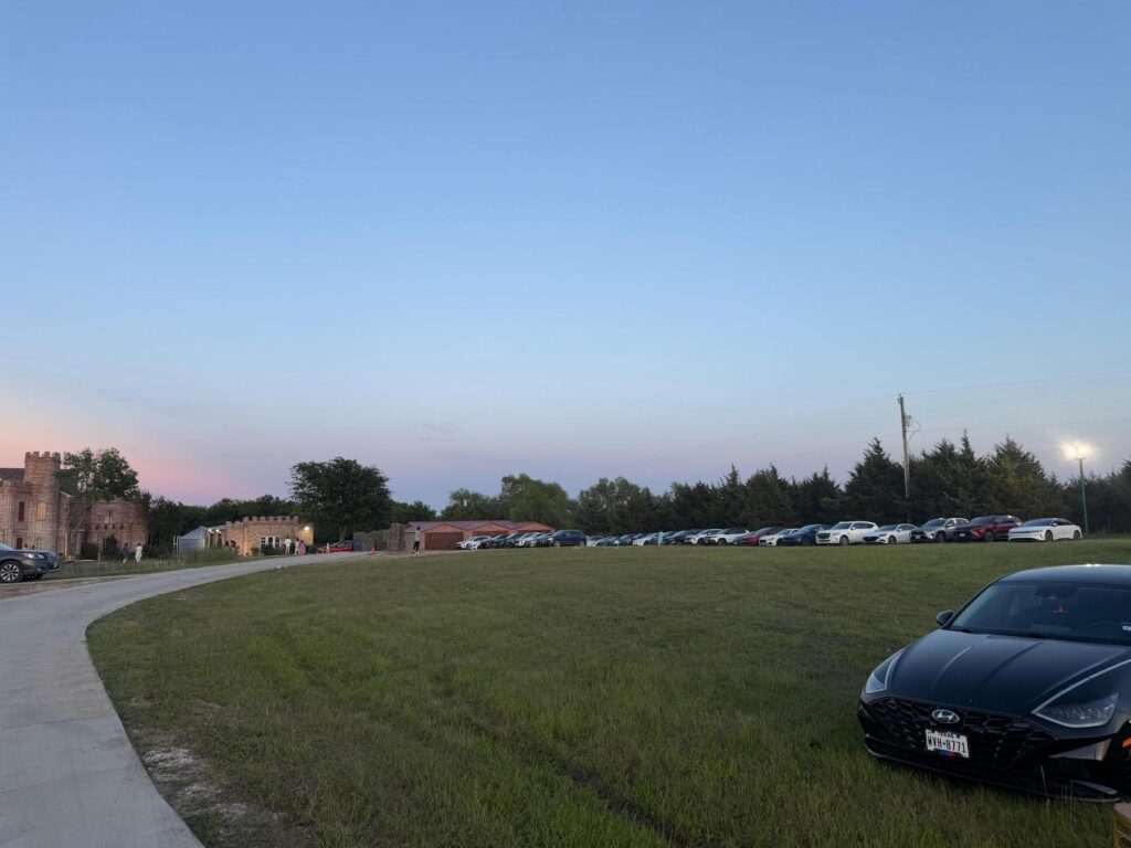 Open grassy field used as parking area with several cars lined up near buildings.