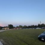 Open grassy field used as parking area with several cars lined up near buildings.