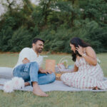 Couple enjoying a romantic outdoor picnic while sitting on a blanket in a green park. The woman is wearing a floral dress, and the couple is smiling and talking beside a picnic basket in a peaceful natural setting.