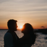 Silhouette of a romantic couple embracing by the water during sunset. The glowing sun is positioned between their faces, creating a warm and intimate atmosphere with soft ocean waves and an orange evening sky in the background.