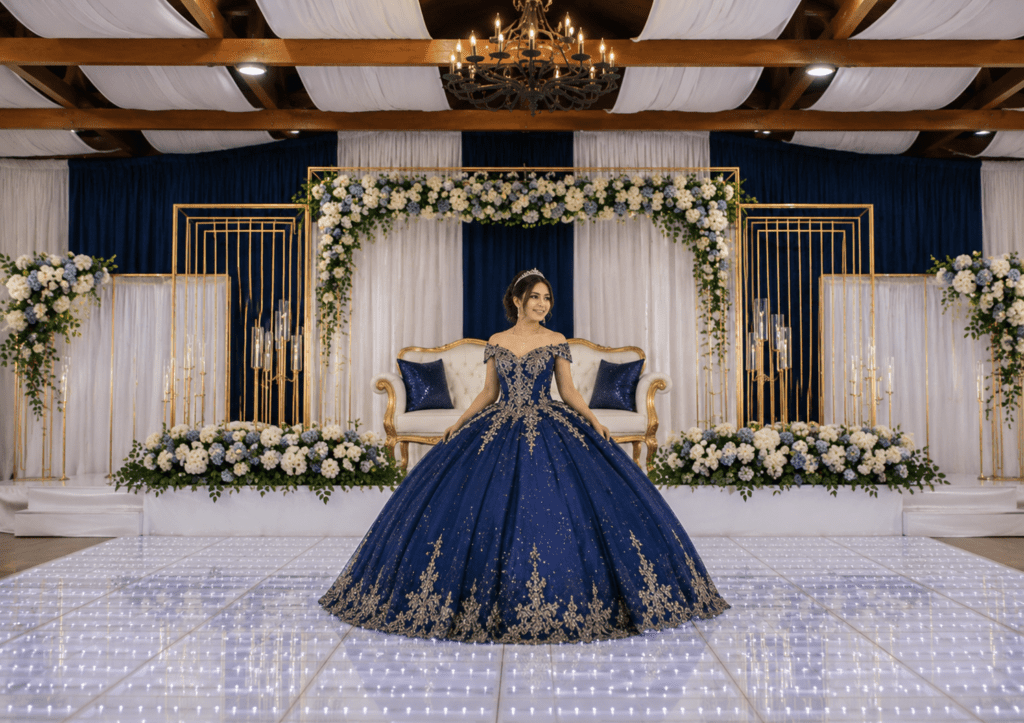 Young woman in a royal blue ball gown standing on a dance floor in front of an elegant floral stage setup during a quinceañera celebration.