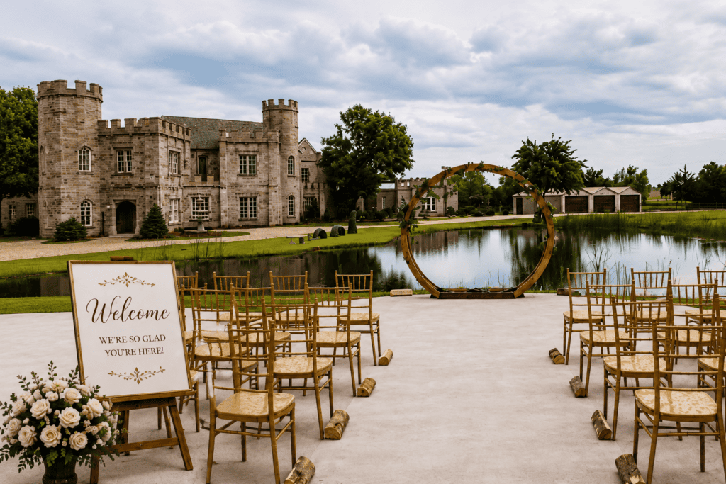 Outdoor wedding ceremony setup with welcome sign, floral arrangement, pond, and castle venue